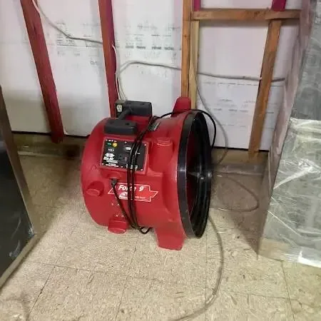Red air mover drying a room with the lower wall cut open and plastic sheeting hung after a floor leak
